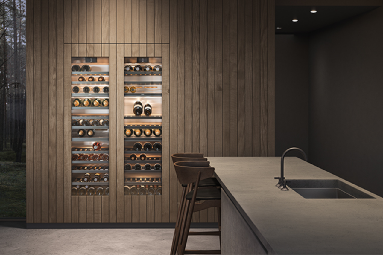 Een sfeervol beeld van een keuken in hout, met een eiland met spoelgedeelte op de voorgrond en twee wijnklimaatkasten van de Gaggenau 400 series, naadloos geïntegreerd in de achterwand van de keuken.