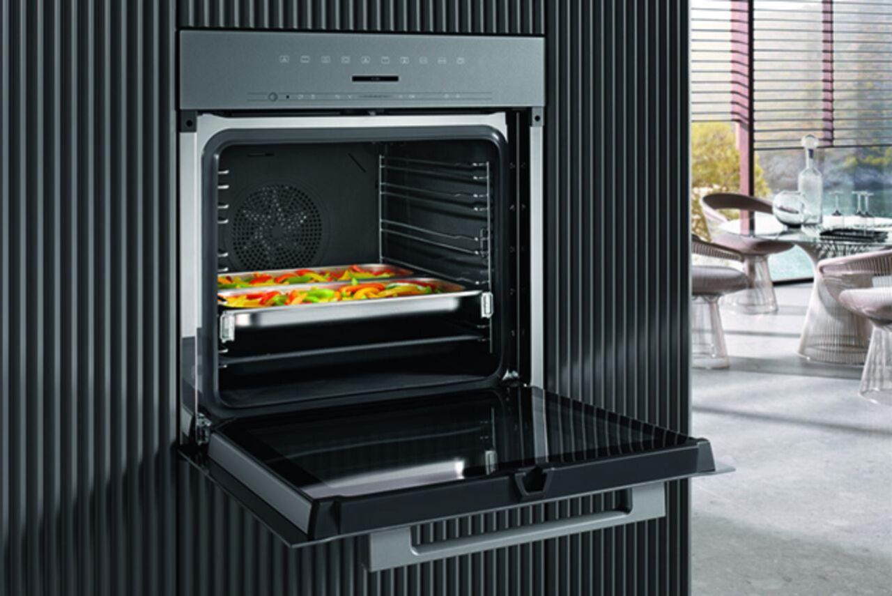 Een nieuwe oven van Miele in de keukenwand van een hedendaags interieur. De deur staat open, zodat je de bakplaat gevuld met veelkleurige paprika's kan zien.
