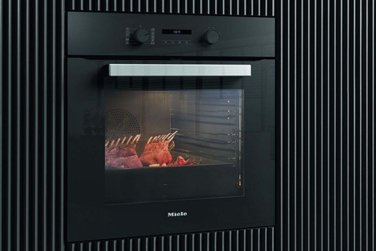 Je ziet een gesloten oven van Miele geïntegreerd in een donkere keukenwand en voorzien van twee bedieningsknoppen. De oven is in werking en bereidt een gerecht met lamskroon.