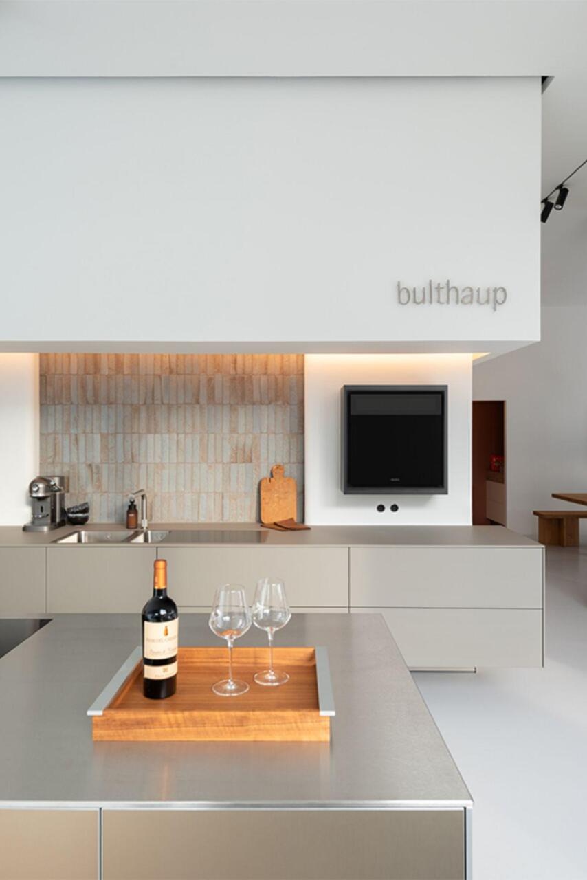 Grijze keuken in de showroom in Gent met fronten en werkbladen in laminaat kleur Flint. Met zwevende wandkasten en spoelbakken op maat ingewerkt in laminaat blad, enkel te zien bij bulthaup. Hoge kasten in massief eiken. Vaatwas op hoogte met push-to-open. Koelkast perfect fresh met push-to-open. Faience verticaal. Quooker kraan.