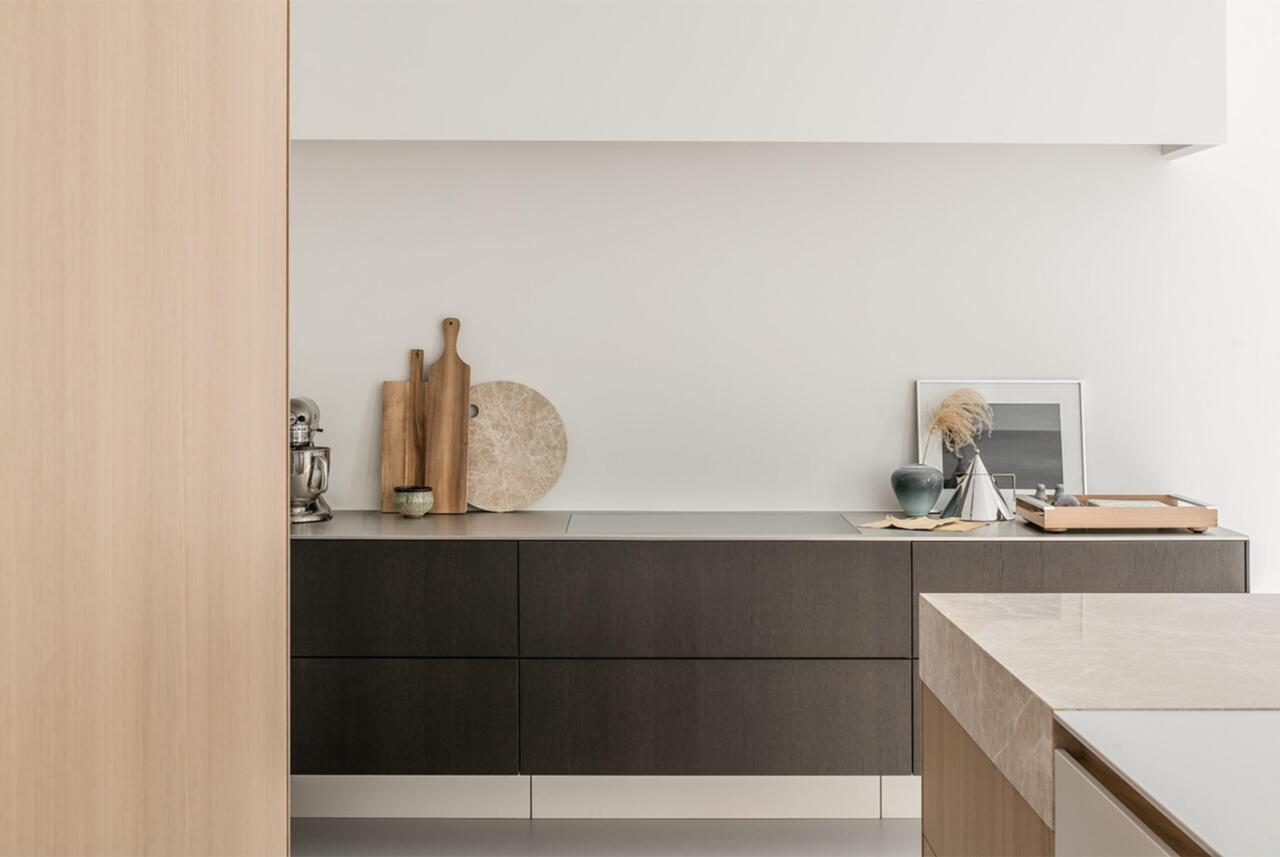 Een frontaal beeld van een deel van een keuken in de showroom met donkerhouten fronten. Links een deel van een hoge kastenwand in bleek hout en rechts een deel van een kookeiland met dik werkblad in beige natuursteen.